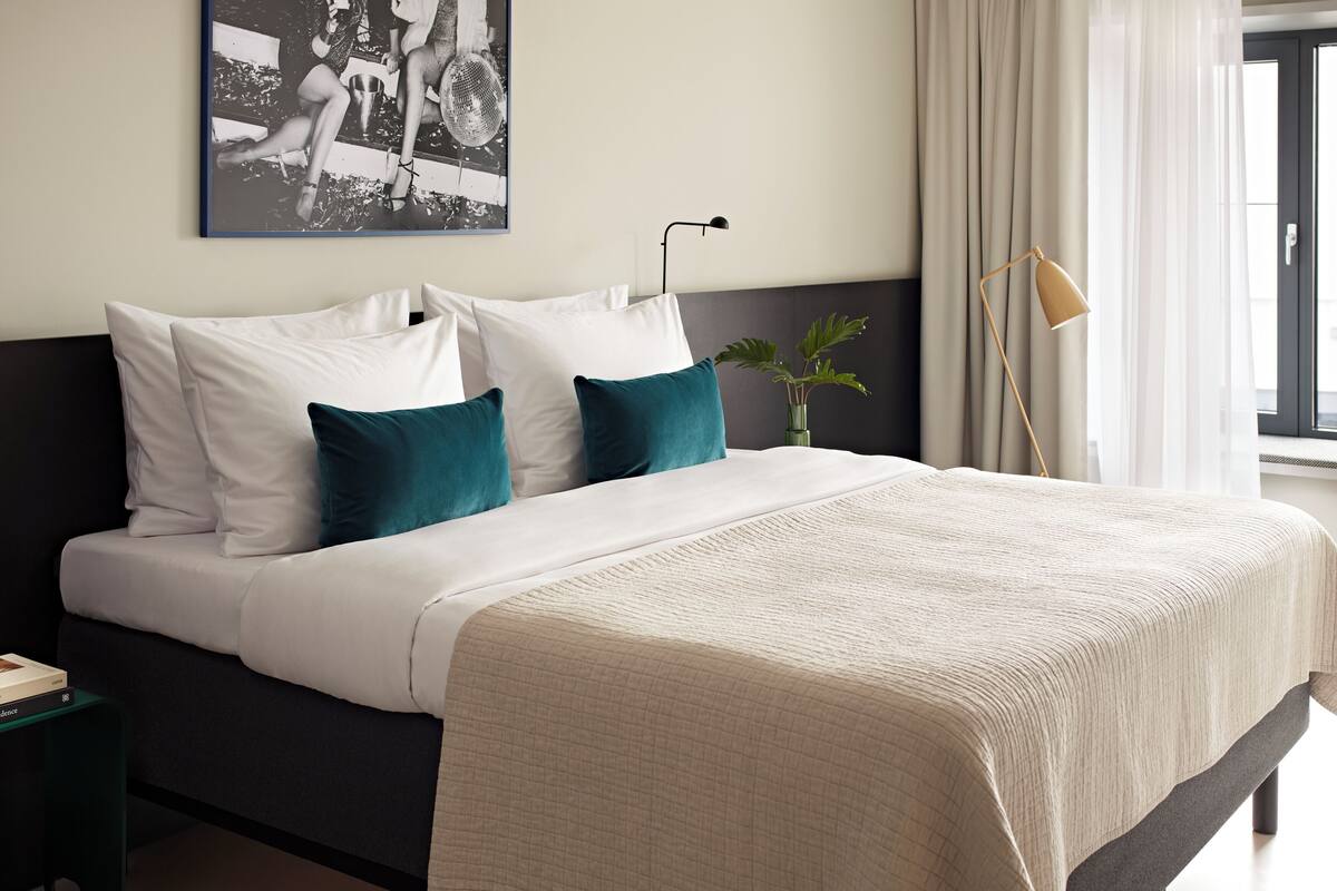 Una cama bien hecha con almohadas blancas y turquesa, una colcha color beige y un cabecero oscuro. Una fotografía en blanco y negro cuelga en la pared sobre la cama.