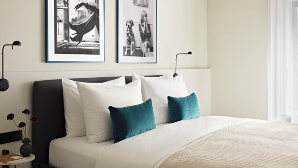 Een moderne slaapkamer met een donker hoofdbord, wit beddengoed, kussens in teal-tinten, en een beige quilt. Twee foto’s in zwarte lijsten hangen aan de muur, een nachtkastje met een bloevas, en een vloerlamp.