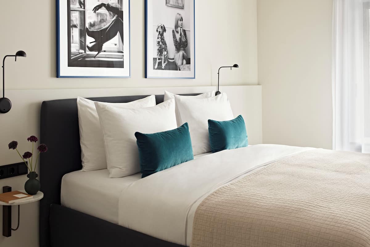 Un dormitorio moderno con cabecera oscura, ropa de cama blanca, almohadas decorativas azul verdoso y una manta acolchada color beige. Hay dos fotografías con marco negro en la pared, una mesita de noche con un jarrón de flores y una lámpara de pie.