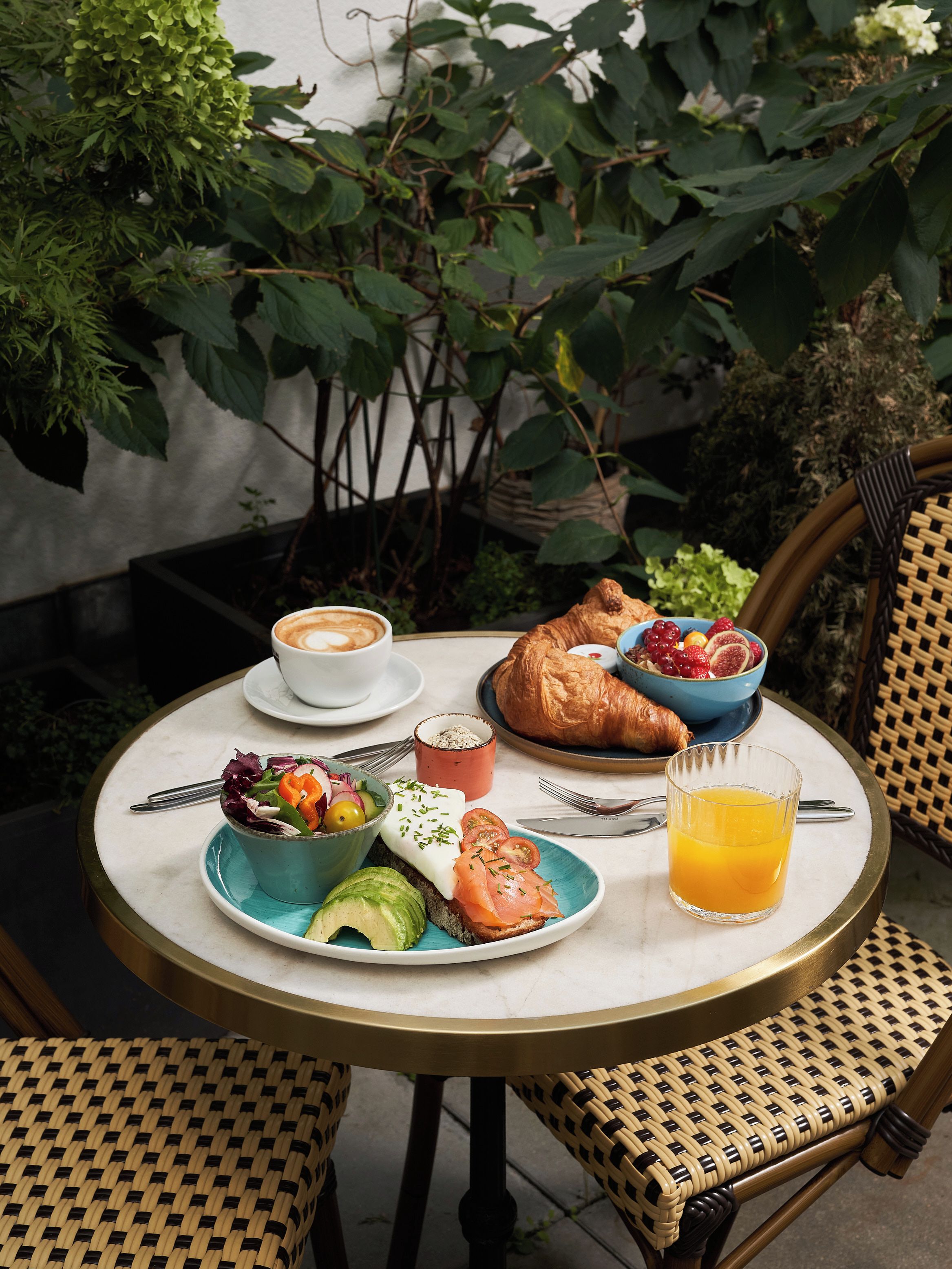 A round table with a white marble top, surrounded by wicker chairs. On the table, there is a plate with a croissant, a bowl of fruit, a glass of orange juice, and a cup of coffee.