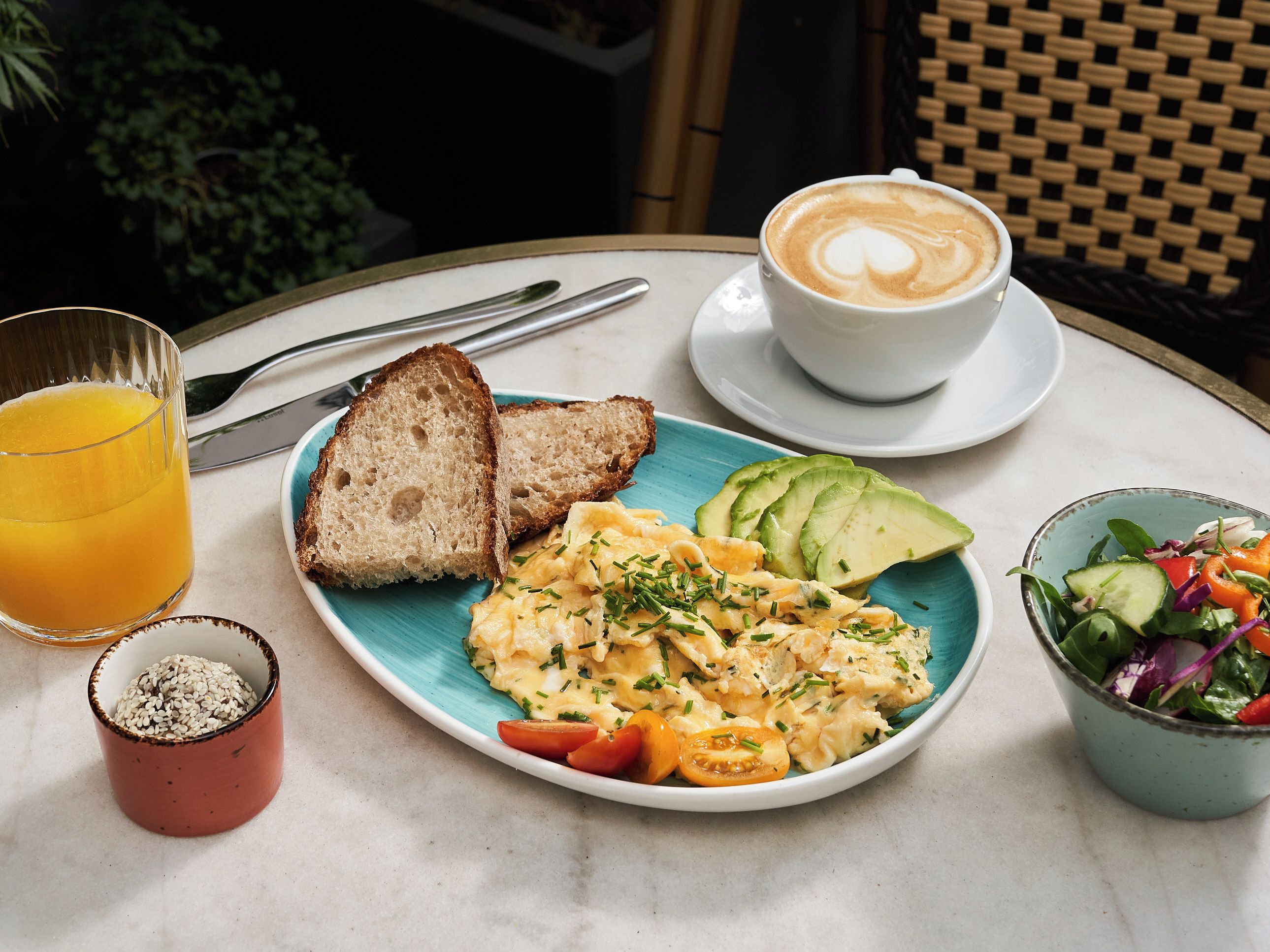 Un desayuno con tostadas, huevos revueltos, aguacate, una ensalada, una taza de café y un vaso de jugo de naranja.