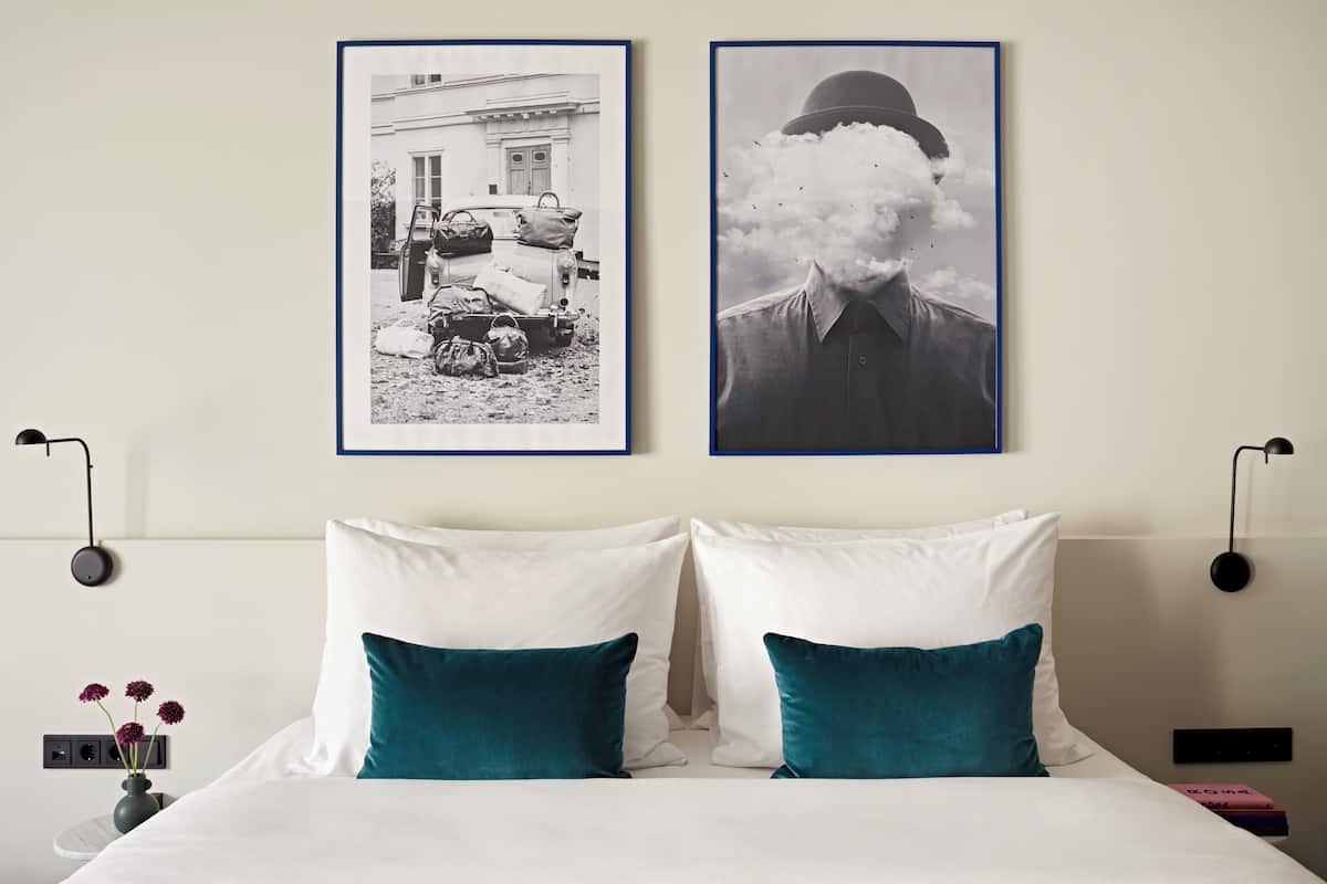 Un dormitorio con una cama, dos almohadas, una mesita de noche, una lámpara y dos cuadros en la pared.