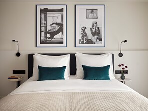 Een net opgemaakt bed met witte en teal kussens, twee zwart-wit foto’s aan de muur, een klein nachtkastje met een bloevas, en een boek op een nachtkastje.