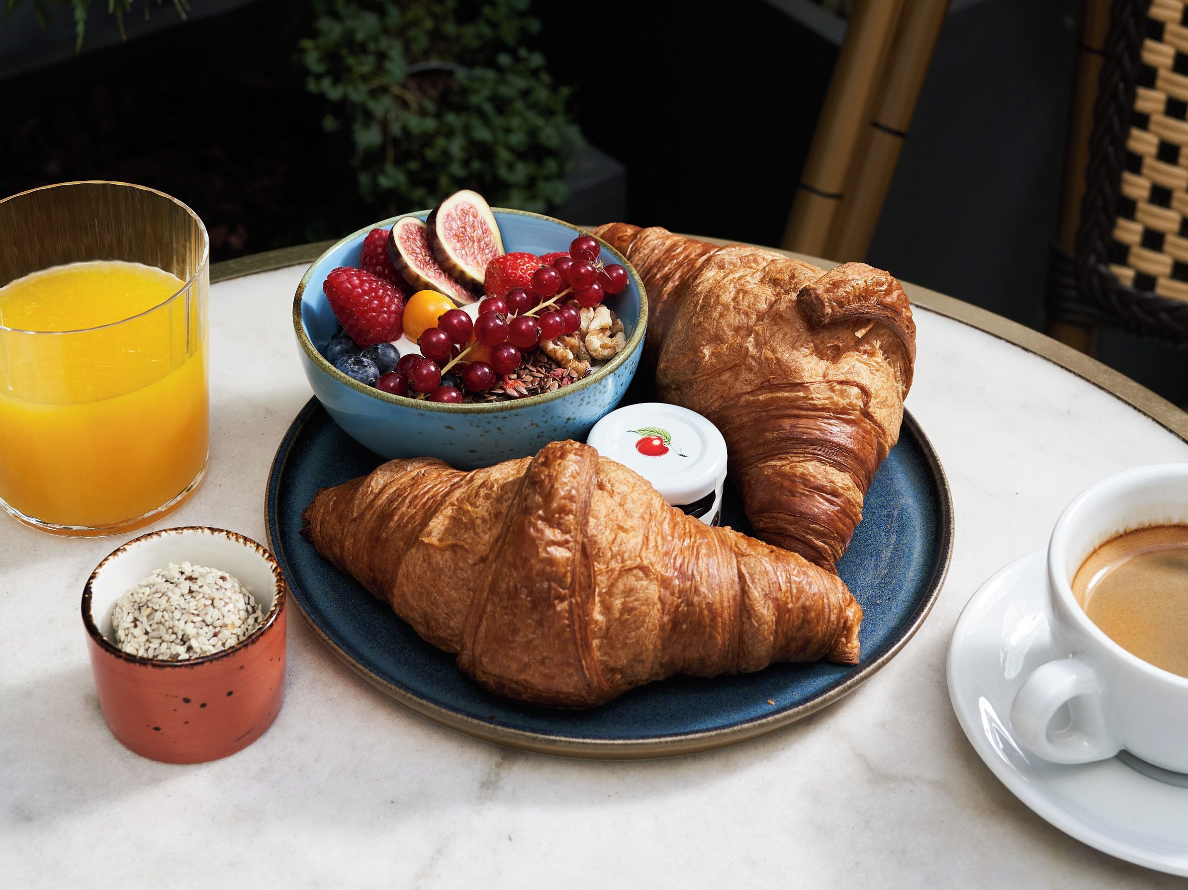 Disposição para o café da manhã com croissants, uma tigela de frutas, um copo de suco de laranja, uma pequena tigela de sementes e uma xícara de café sobre uma mesa.