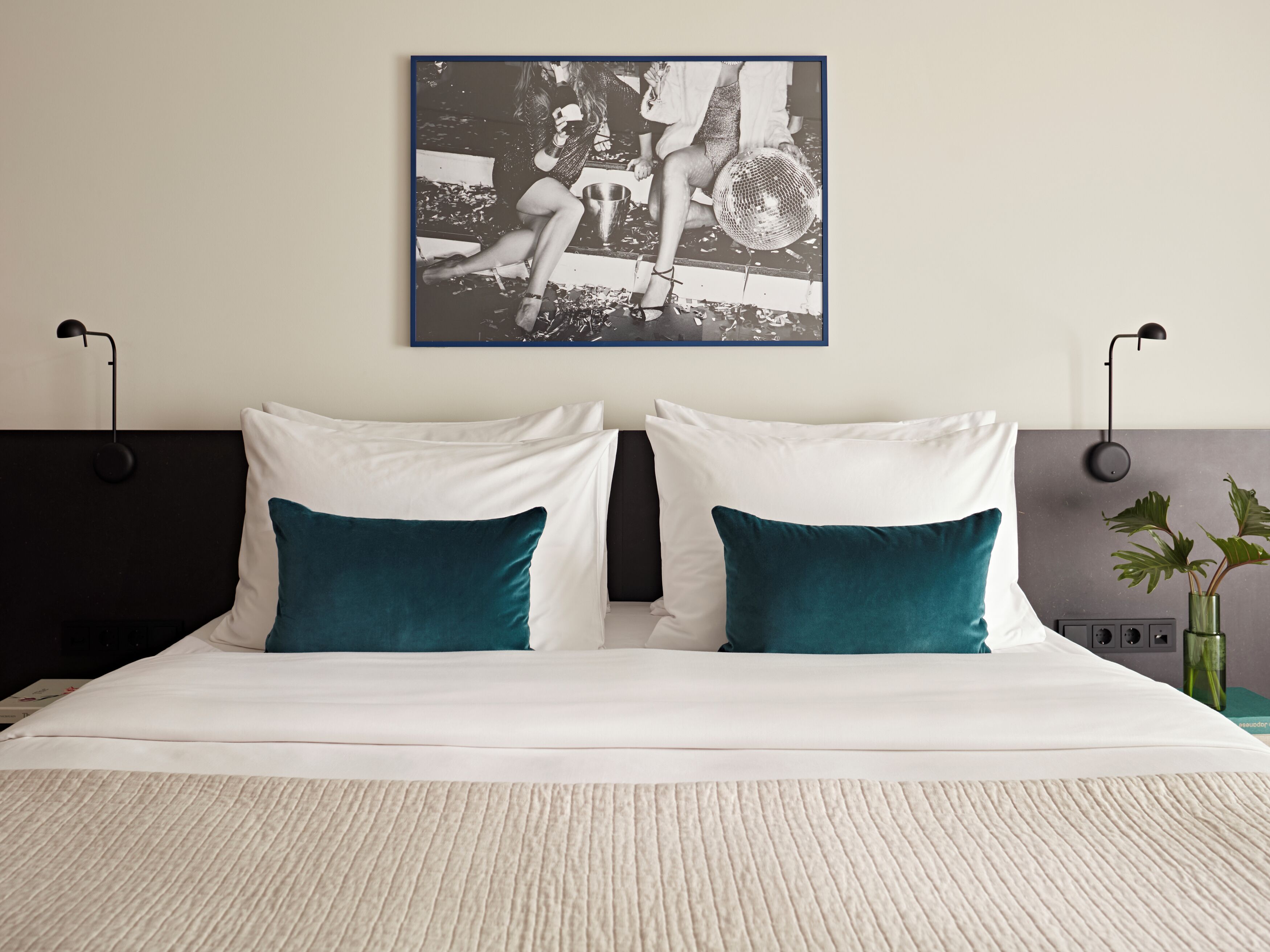 Una cama bien tendida con almohadas blancas y turquesa, una obra de arte blanco y negro encima, y una planta verde en un florero sobre la mesita de noche.
