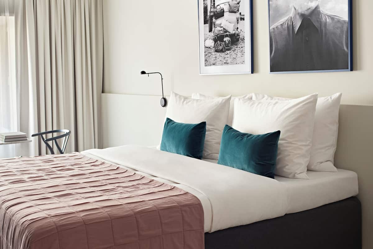 Una cama bien hecha con colcha rosa, almohadas blancas y almohadas de color turquesa. Hay dos cuadros en la pared, una lámpara y una silla en la habitación.