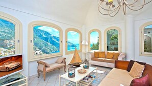 A spacious living room with large windows, a chandelier, a sofa, a coffee table, and a television. - VILLA BIANCA POSITANO (Positano)