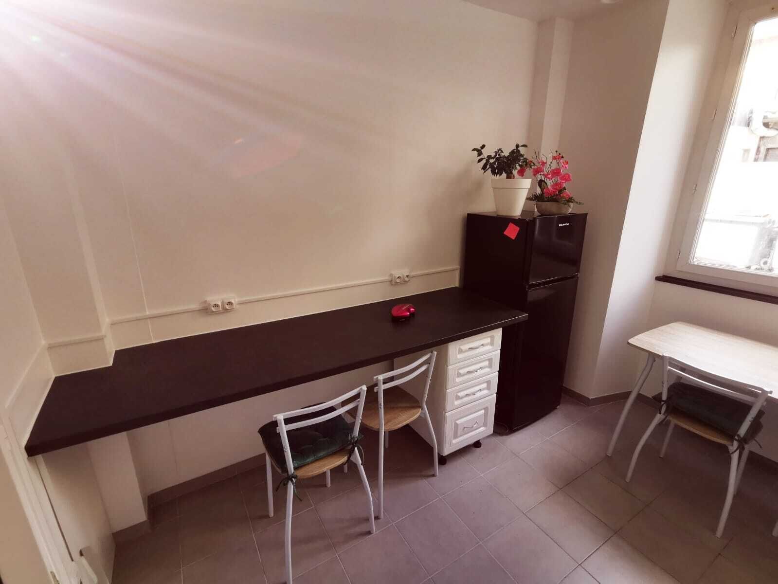 Une petite cuisine équipée avec un plan de travail noir, un réfrigérateur, un évier, deux chaises et une fenêtre.