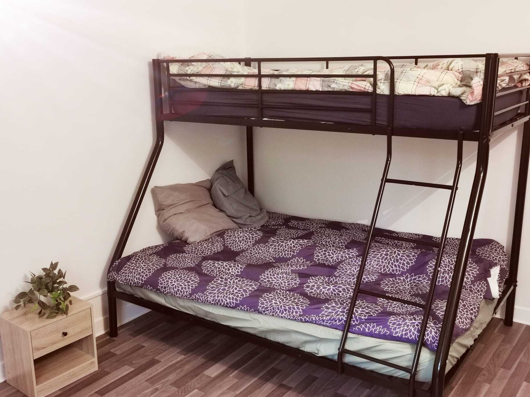 A bunk bed with a purple floral bedspread, a wooden nightstand with a plant, and a white wall.