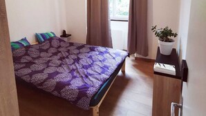 A bedroom with a bed, a window with curtains, a bedside table with a plant, and a door.