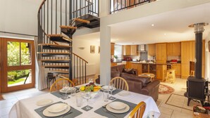 A dining area with a table set for four, a spiral staircase, a kitchen with wooden cabinets, and a wood-burning stove. - Harthanger View Cottage, Luxborough (Wiveliscombe)