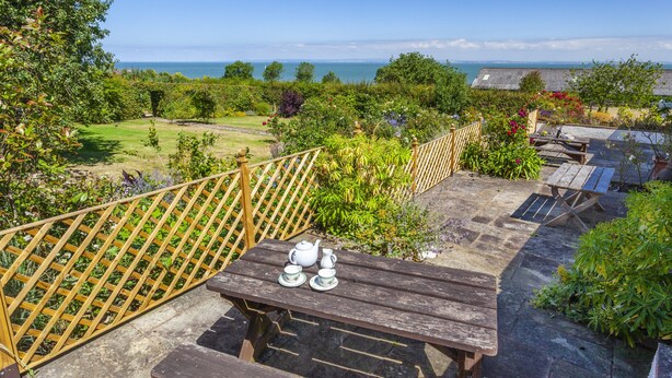 Una terraza de madera con una mesa dispuesta para el té, rodeada de abundante vegetación y vistas al mar.