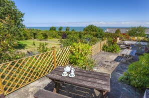 A wooden deck with a table set for tea, surrounded by lush greenery and a view of the sea. - Coach House View, Porlock Weir (Porlock Weir)