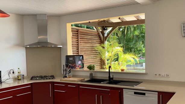 A modern kitchen with red cabinets, a stainless steel range hood, a window with a view of greenery, and a dishwasher. - come and relax at CASA FALL, 8 minutes from the DIDIER waterfalls, by the pool (Schoelcher)
