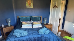 Un dormitorio con colcha azul acolchada, dos almohadas con estampado de paisaje, cabecero de madera, lámparas de noche y una puerta con perchero.