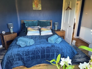A bedroom with a blue quilted bedspread, two pillows with a landscape print, a wooden headboard, bedside lamps, and a door with a hanging coat rack. - Charming rural cottage, with stove & large garden overlooking Loch Awe, Argyll. (Kilchrenan, by Taynuilt)