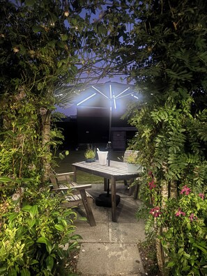 An outdoor patio area with a table and chairs, surrounded by lush greenery and a decorative umbrella. - Charming rural cottage, with stove & large garden overlooking Loch Awe, Argyll. (Kilchrenan, by Taynuilt)