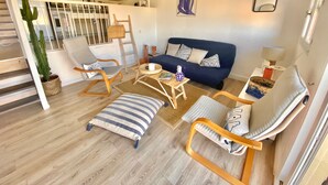 Een gezellige woonkamer met een blauwe bank, houten stoelen, een gestreept poef en een cactus.