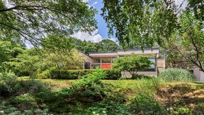Een modern huis met twee verdiepingen en een plat dak, omringd door weelderige begroeiing en bomen.