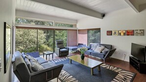 Een moderne woonkamer met een bank, fauteuils, een salontafel en een televisie. Grote ramen bieden uitzicht op de omgeving.