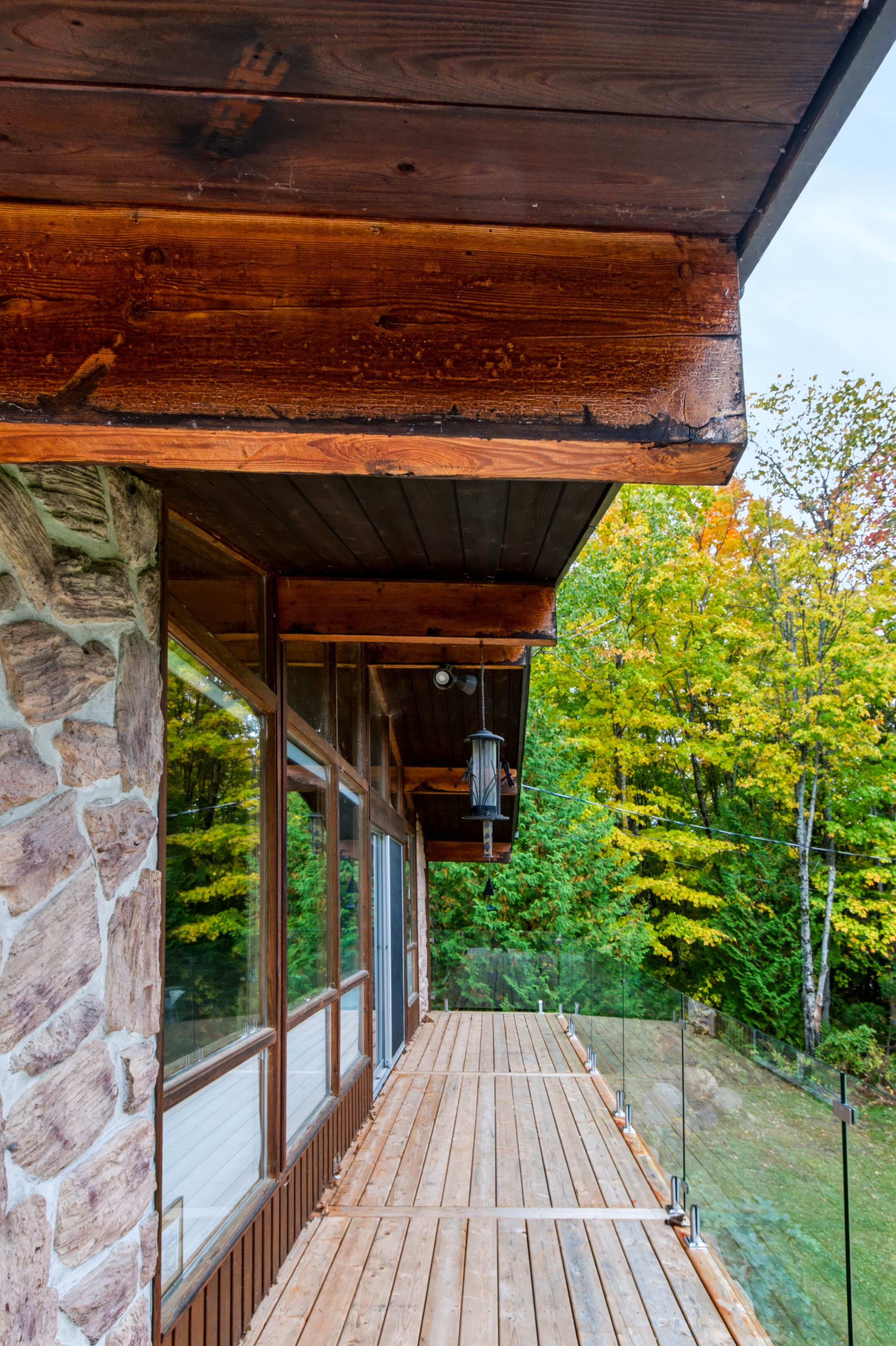 Une terrasse en bois, avec un mur en pierre d’un côté et une porte vitrée donnant sur un porche abrité.