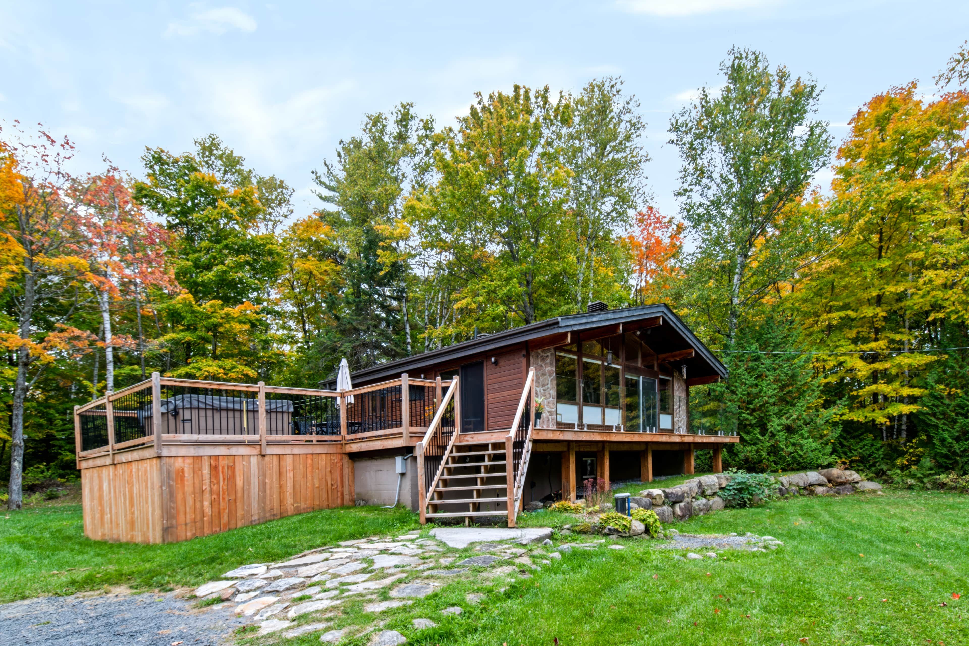 A cabin with a wooden deck, a stone staircase, and a view of a forest with autumn foliage.