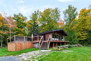 A cabin with a wooden deck, a stone staircase, and a view of a forest with autumn foliage.