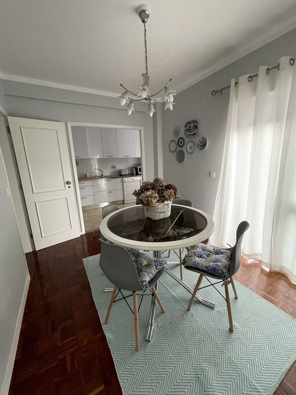 A dining area with a round table, chairs, a chandelier, and a kitchen in the background.