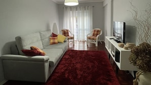 Een woonkamer met een grijze bank, een rood kleed en een televisie op een witte dressoir.