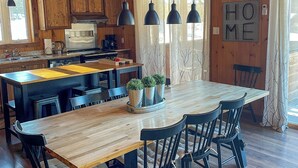 Un interior de madera con una gran mesa de comedor, sillas negras, lámparas colgantes y una zona de cocina con armarios de madera y un refrigerador.