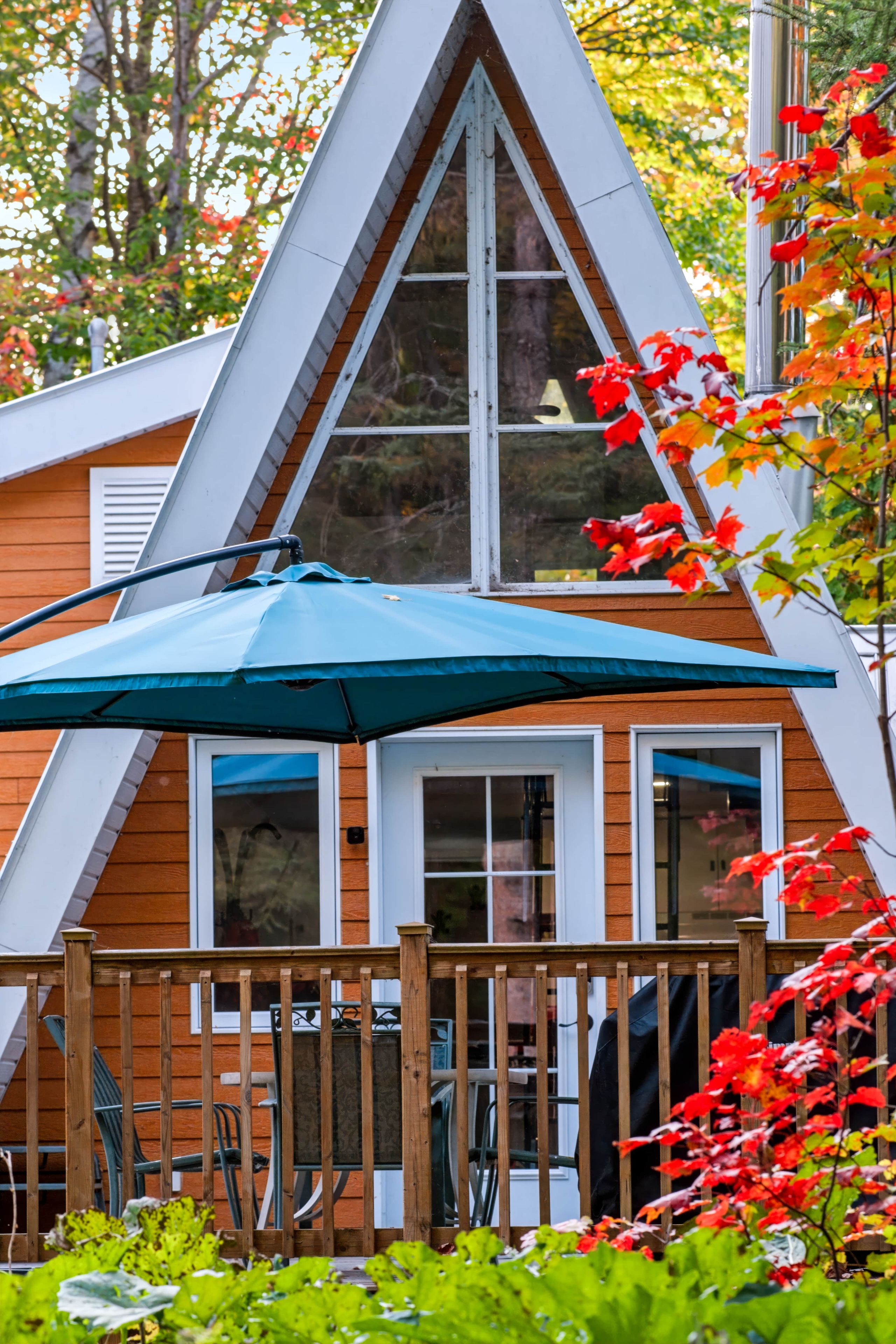 Une cabane en bois avec un toit triangulaire, une terrasse dotée d’un parasol bleu et une clôture en bois.