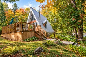 A cabin with a wooden deck, stairs, and a metal roof, surrounded by autumn foliage.
