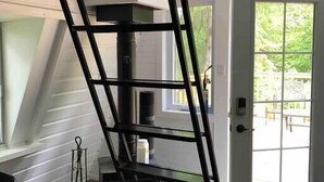 A room with a black metal ladder leaning against a white wall, a fireplace, and a door leading to an outdoor area.
