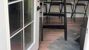 A room with a sliding glass door, a black metal ladder, a dining table with chairs, and a kitchen area with cabinets and appliances.