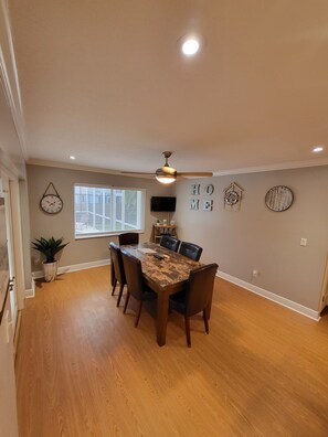 Un coin repas comprenant une table en bois, six chaises, un ventilateur de plafond et une horloge murale.