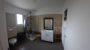 A bathroom with a glass shower enclosure, a white sink, a mirror, and a wooden stool. - Maison au Calme (Aléria)