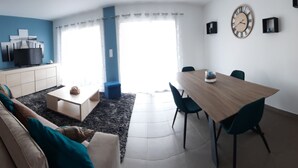 A modern living room with a sofa, coffee table, and dining table. There is a TV on a cabinet, a clock on the wall, and a window with curtains. - Maison au Calme (Aléria)