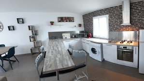 A modern kitchen with a brick wall, a dining area with a long table and chairs, and a built-in washing machine. - Maison au Calme (Aléria)