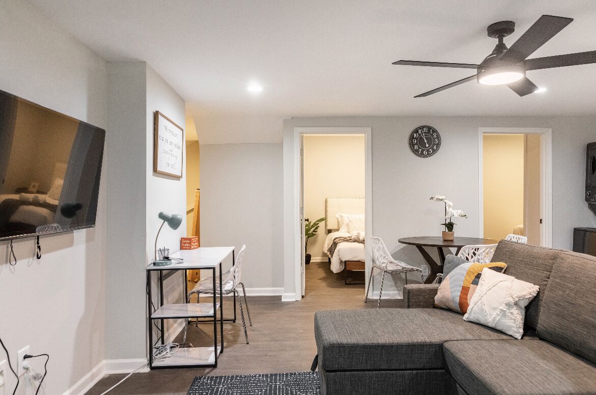 A modern living room with a gray sofa, a dining table with chairs, a ceiling fan, and a TV mounted on the wall.