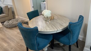 Un área de comedor con una mesa redonda, cuatro sillas tapizadas en azul, un florero con flores blancas y un pequeño adorno sobre la mesa.