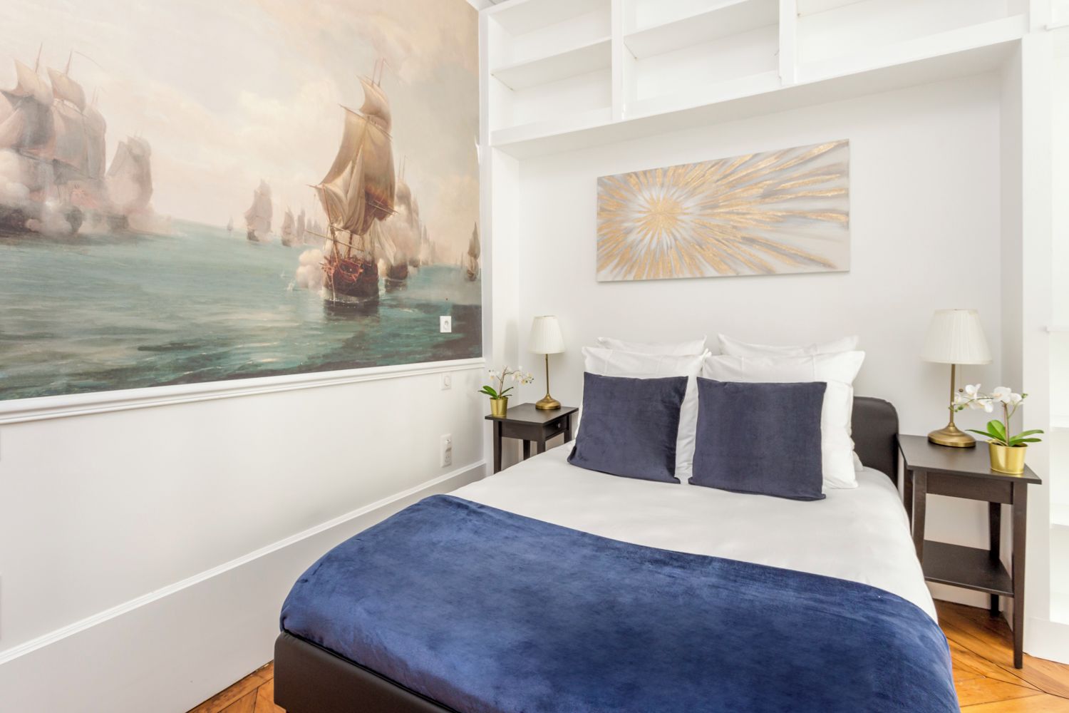 A modern hotel room with a large bed, two bedside tables with lamps, a large abstract painting, and a view of ships on the water.