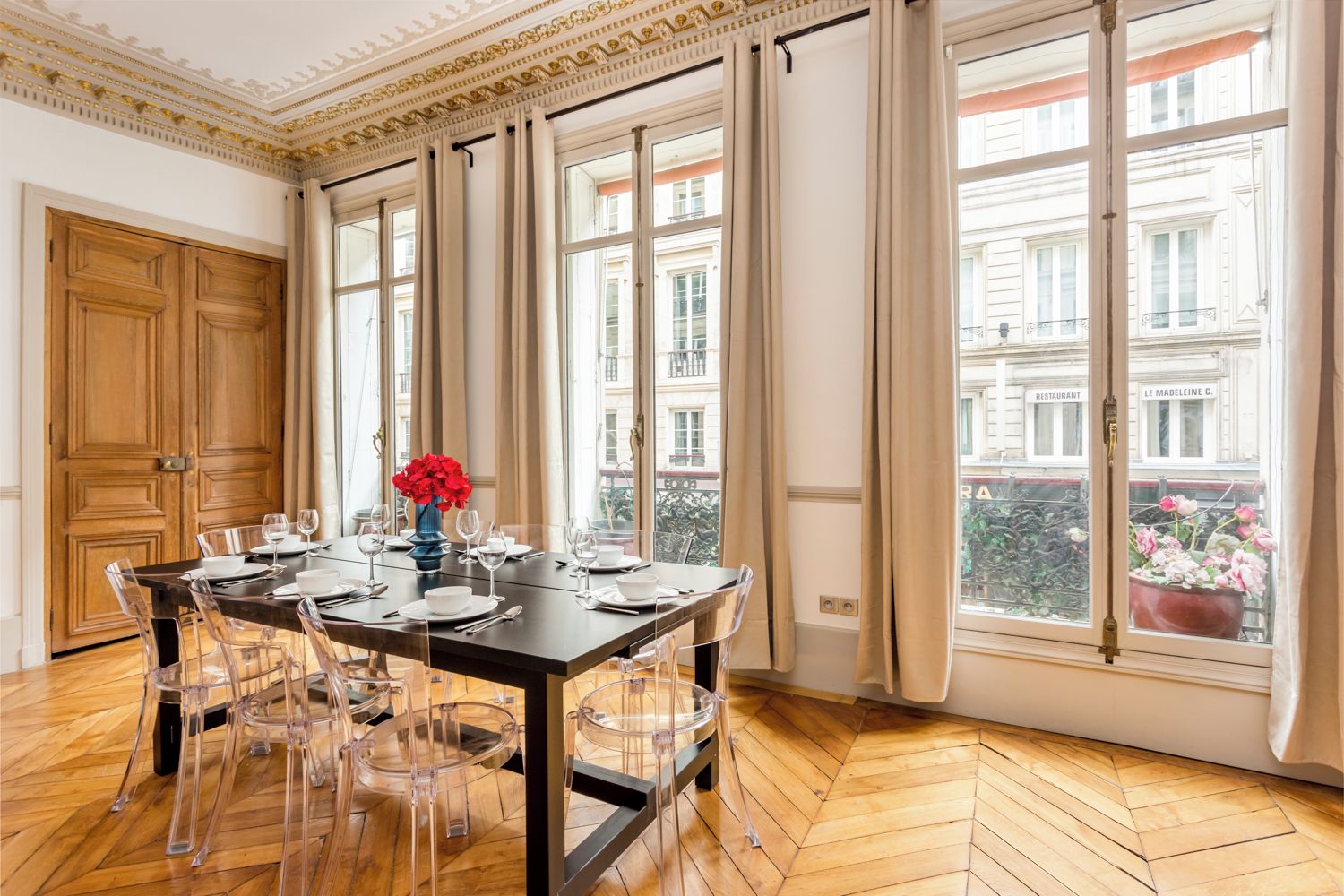Ein Esszimmer mit einem großen Tisch, gedeckten Plätzen für sechs Personen, transparenten Stühlen und einer Vase mit roten Blumen.