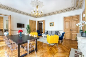 Un vaste salon doté d’un parquet en bois disposé en point de Hongrie, d’une table à manger noire avec des chaises transparentes, d’un canapé et d’un téléviseur fixé au mur.