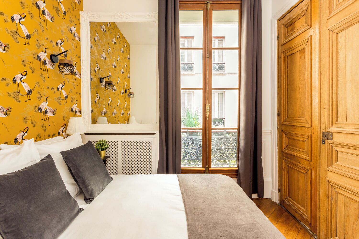 Una camera da letto con un letto, uno specchio, una finestra vista edifici e una porta in legno.