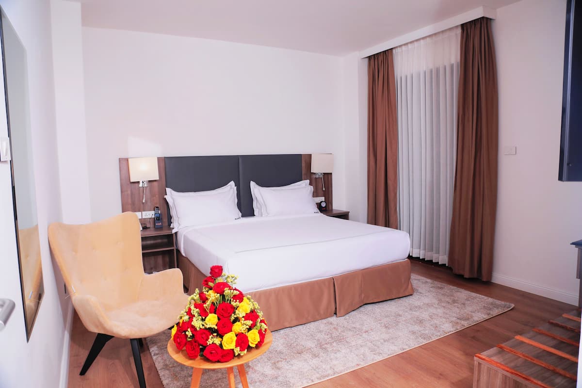 A hotel room with a large bed, a chair, a nightstand, a lamp, a bouquet of flowers, and a window with curtains.