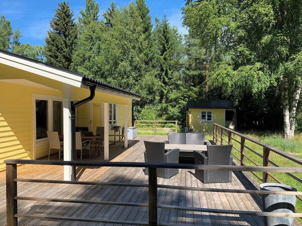 A wooden deck with outdoor furniture, a yellow building with a covered patio, and a forested background.