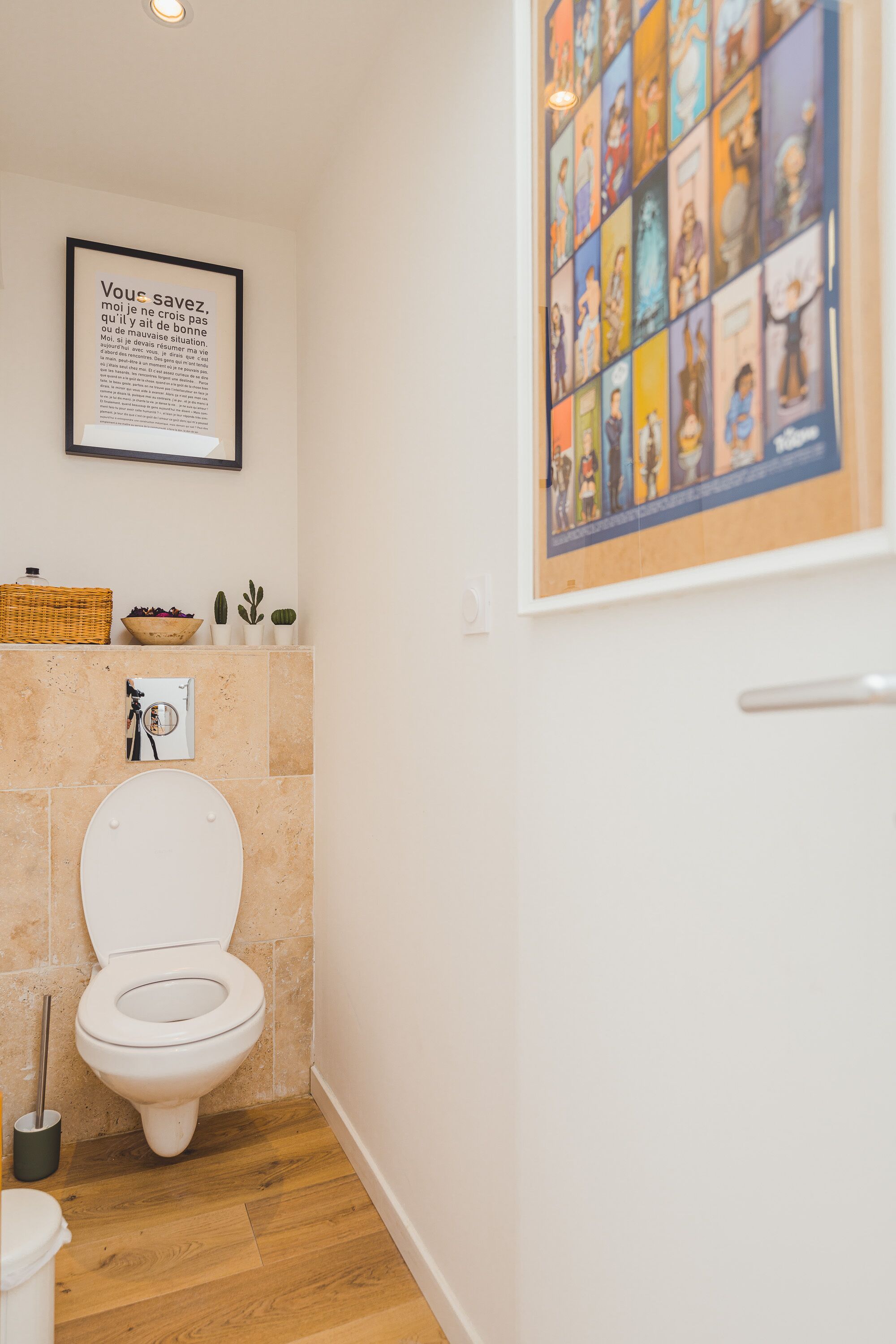 Een badkamer met een toilet, een kaderrand citaat aan de muur, een klein plankje met planten en een poster met tekenfilmfiguren.