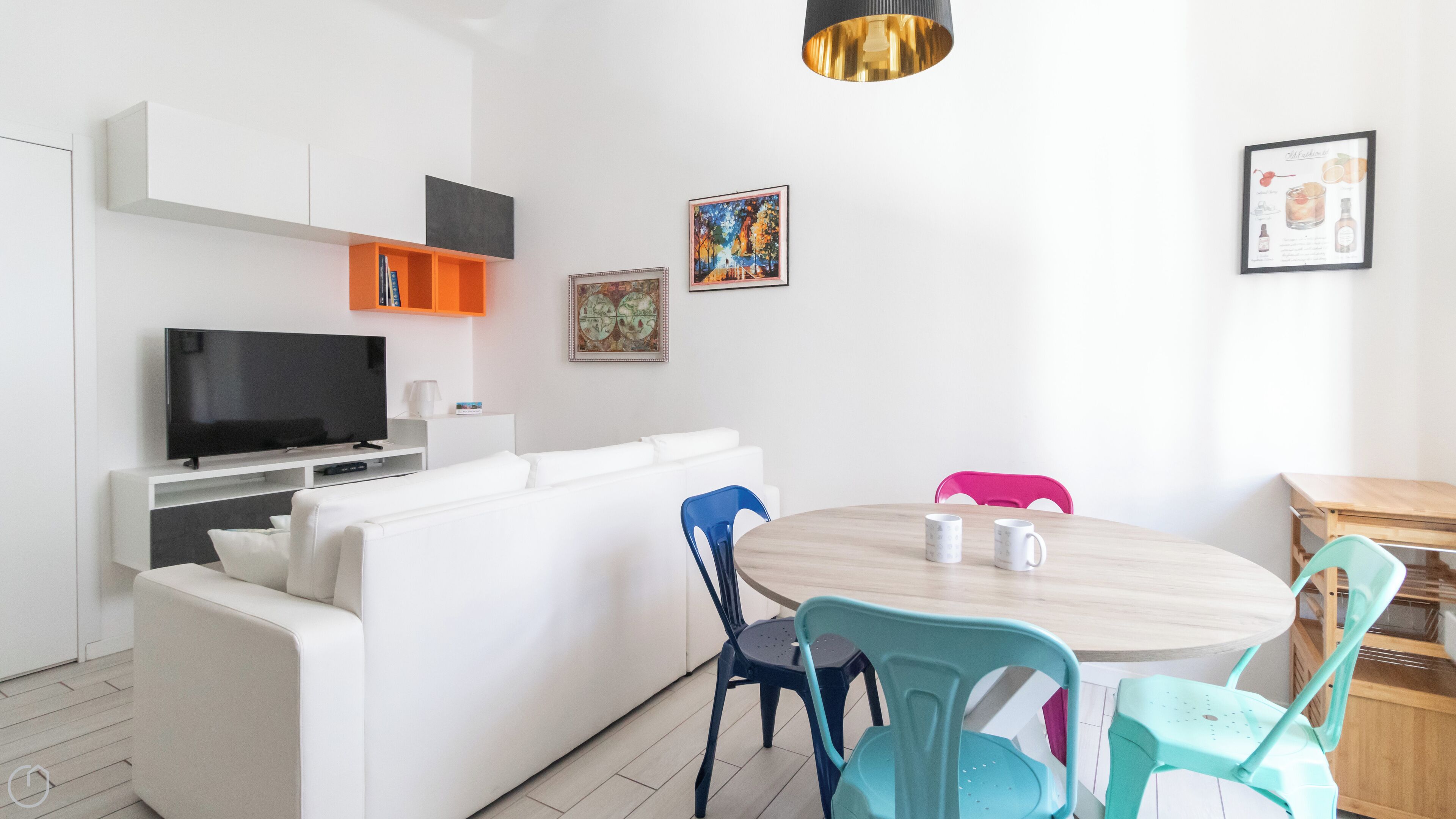 A modern living room with a white sofa, a TV, a dining table with chairs, and artwork on the walls.