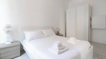 A minimalist white bedroom with a bed, nightstands, a wardrobe, and neatly folded towels.