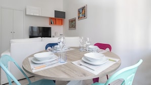 A dining area with a round wooden table set for four, white plates, and clear glasses. There are two blue chairs and one pink chair. A white sofa and a TV are in the background. - Italianway Easy - Arbe 51 (Milan)